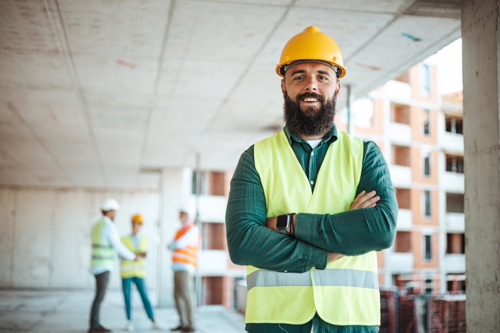 Construction worker in a yellow safety vest and hard hat standing confidently on site with a modern building in the background.