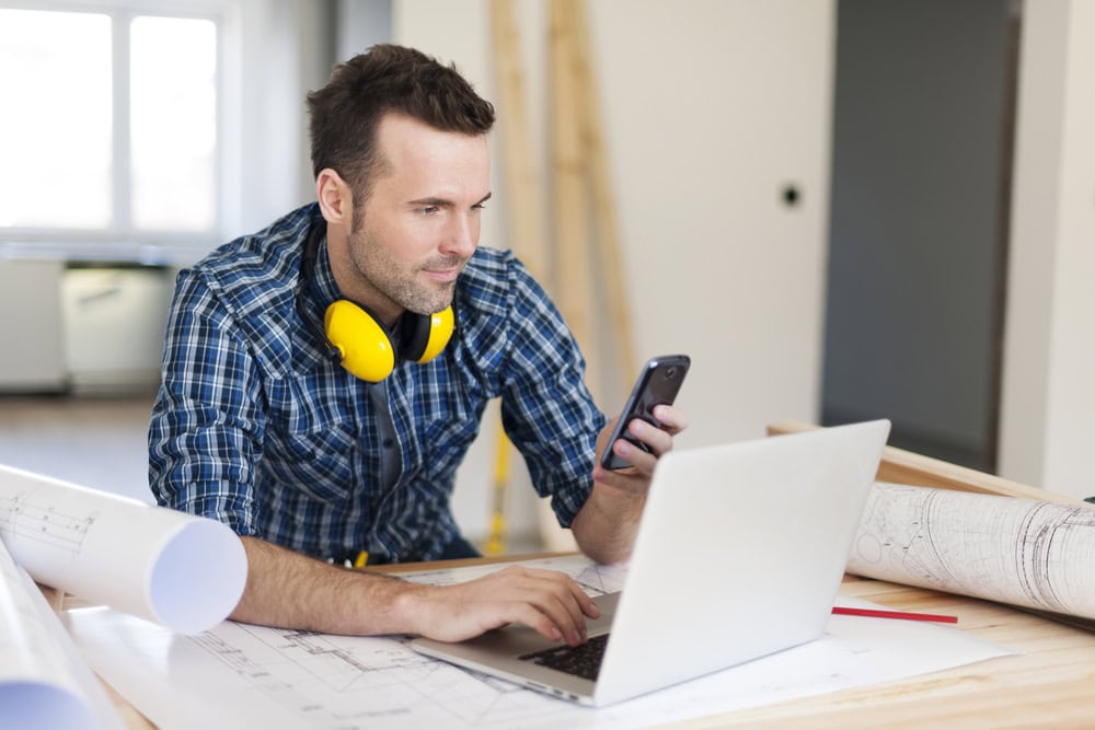 Hard hat and blue plaid shirt wearing man working on a construction project, using a smartphone and laptop at a worksite.