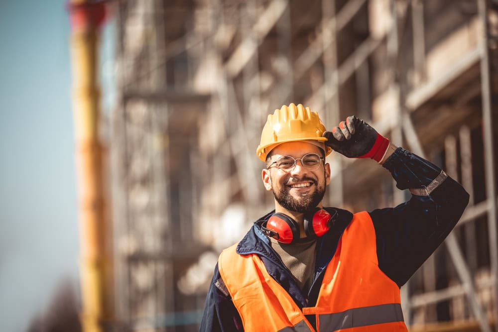 Construction worker smiling in hard hat and safety gear at building site.