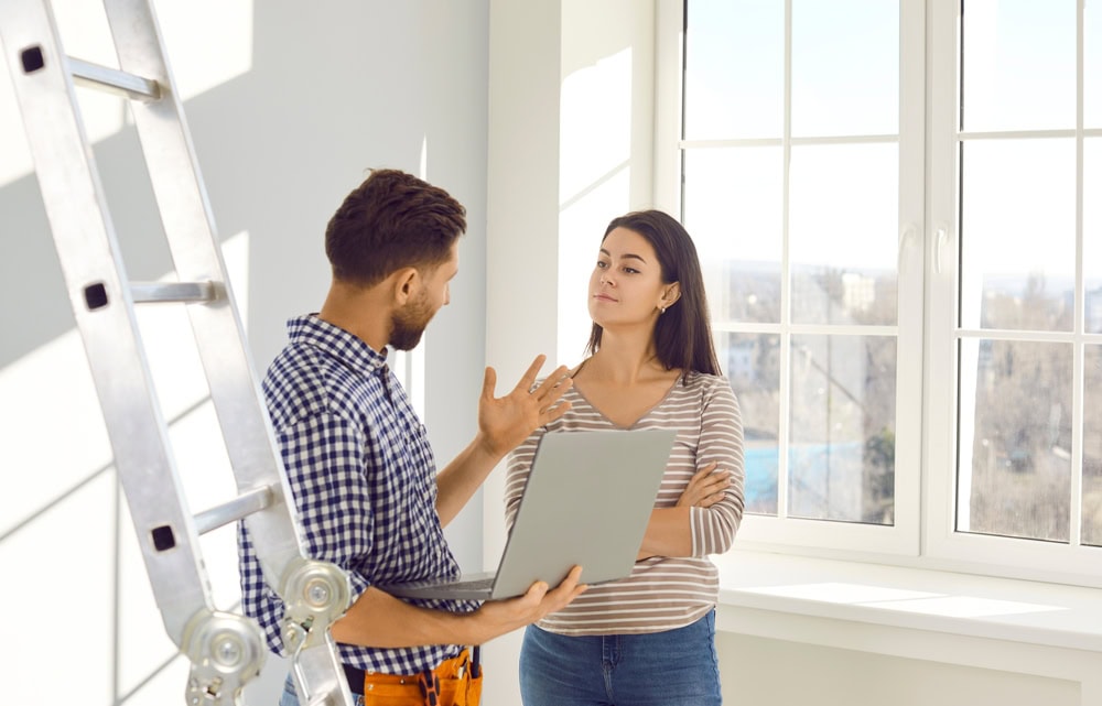 Overgrown contractor with a ladder discussing project details with a female client in a well-lit room.