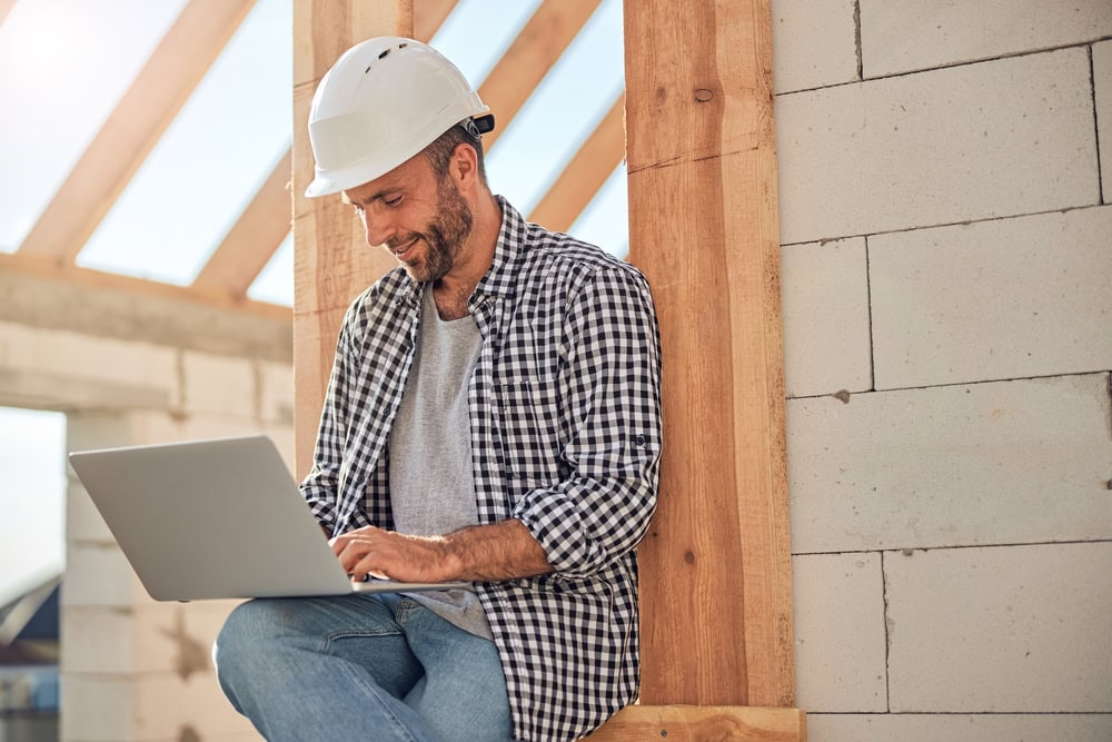 Male construction worker with hard hat using a laptop at a construction site.