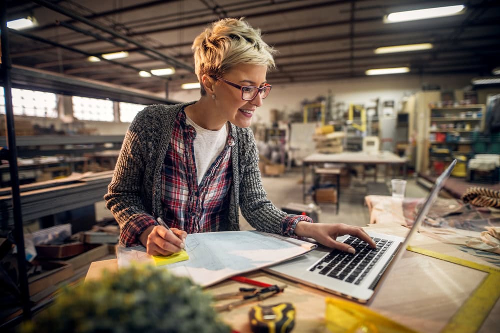 Experienced woman in a woodworking shop working on laptop, preparing for contractor licensing or business planning.