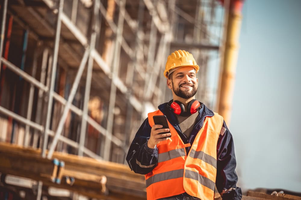 Construction worker in a hard hat and safety vest on a building site, smiling and using a smartphone.