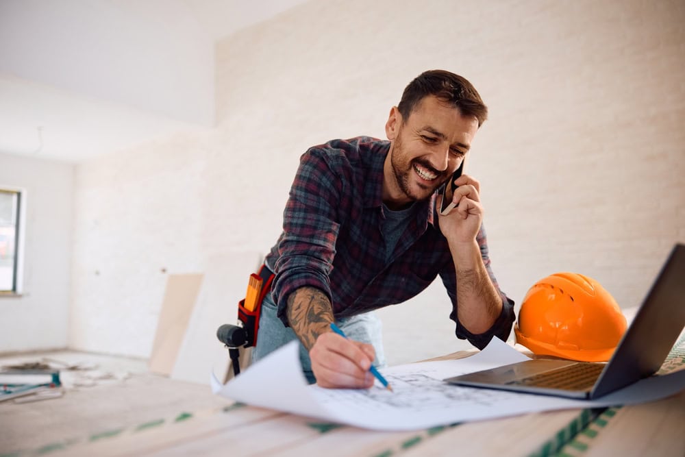 Friendly male contractor working with blueprints, talking on his phone, in an unfinished interior space, wearing a tool belt and safety helmet.