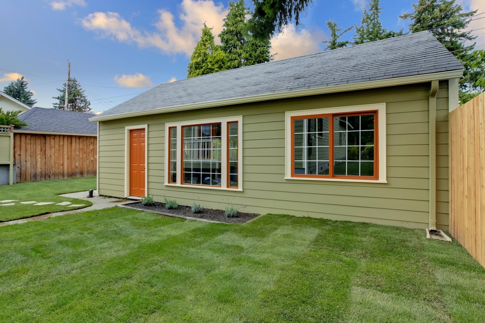 Bright green backyard with a newly painted small house featuring orange window frames and a single door, surrounded by a wooden fence and lush grass, under a partly cloudy sky.