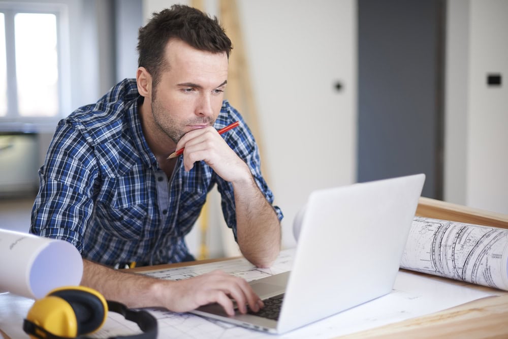 Focused man working on blueprints at a desk with a laptop, symbolizing contractor planning and preparation.