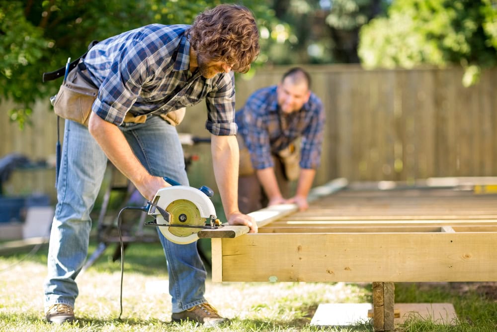 High school graduate installing wooden deck using circular saw in backyard.