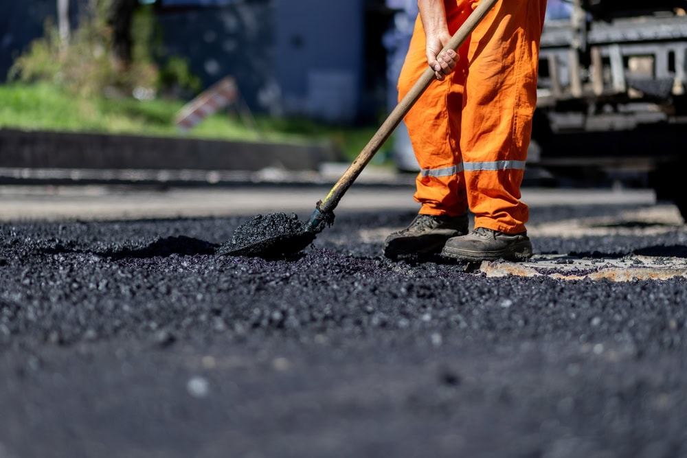A construction worker in orange safety gear spreading fresh asphalt on a road surface with a shovel.