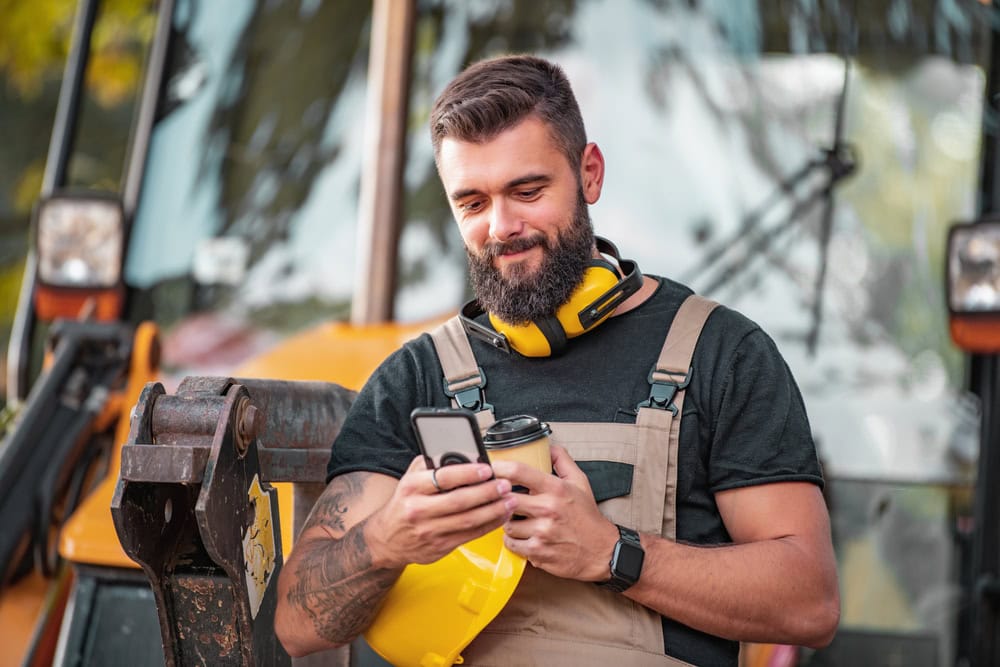 A skilled construction worker checking his phone at a construction site with heavy equipment in the background.
