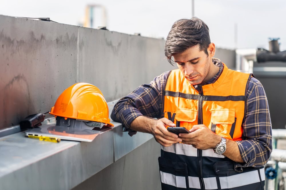 Hard hat, tools, and construction plans on rooftops with a young male contractor using a smartphone.