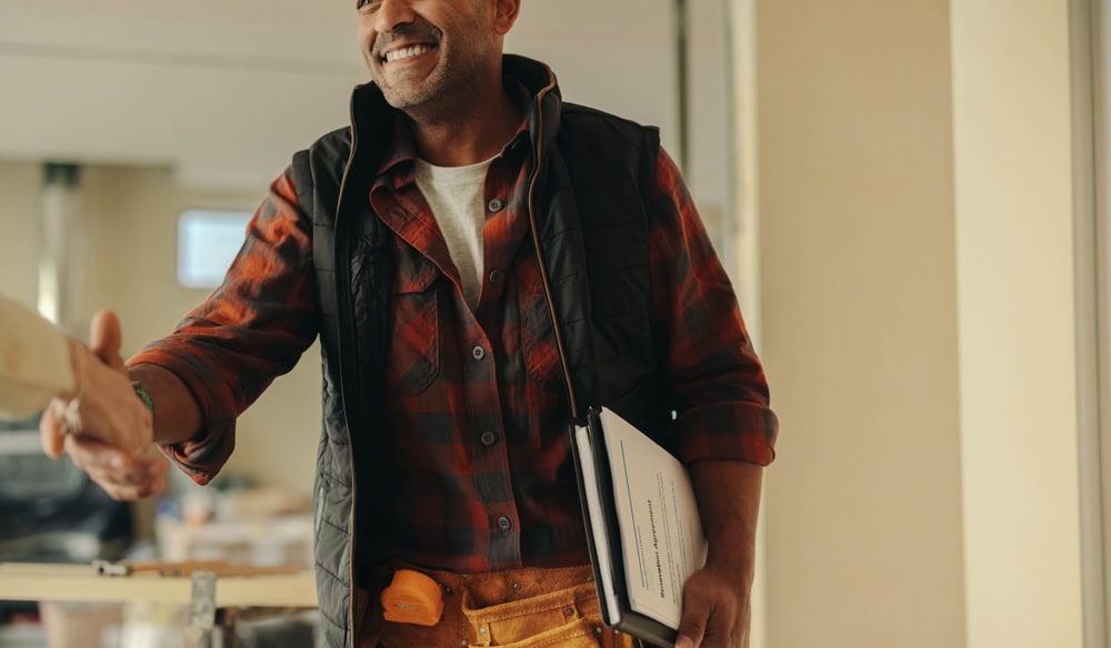 Friendly contractor shaking hands with client indoors, holding a clipboard and smiling confidently, symbolizing trust and professionalism in the construction industry.