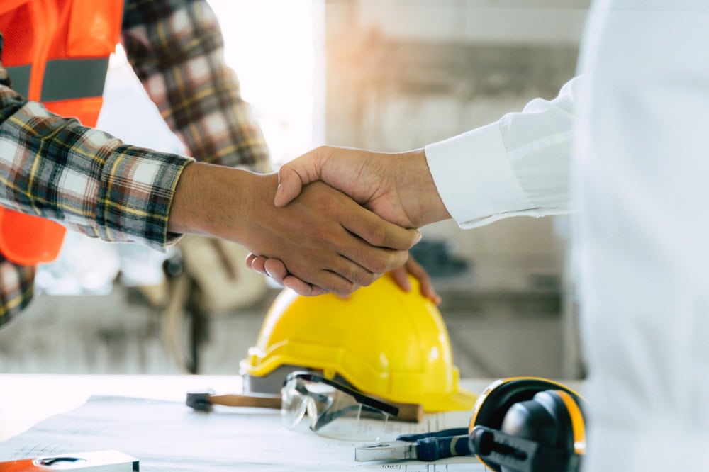 Two workers shaking hands, with construction helmets and tools on a table, symbolizing partnership in the construction industry. | Construction handshake between workers in a project site.