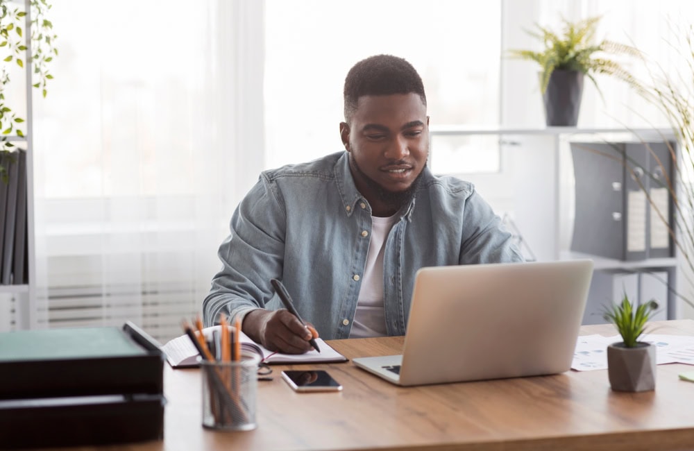 Young man working on a laptop at a desk with office supplies and plants, studying for contractor licensing exam.