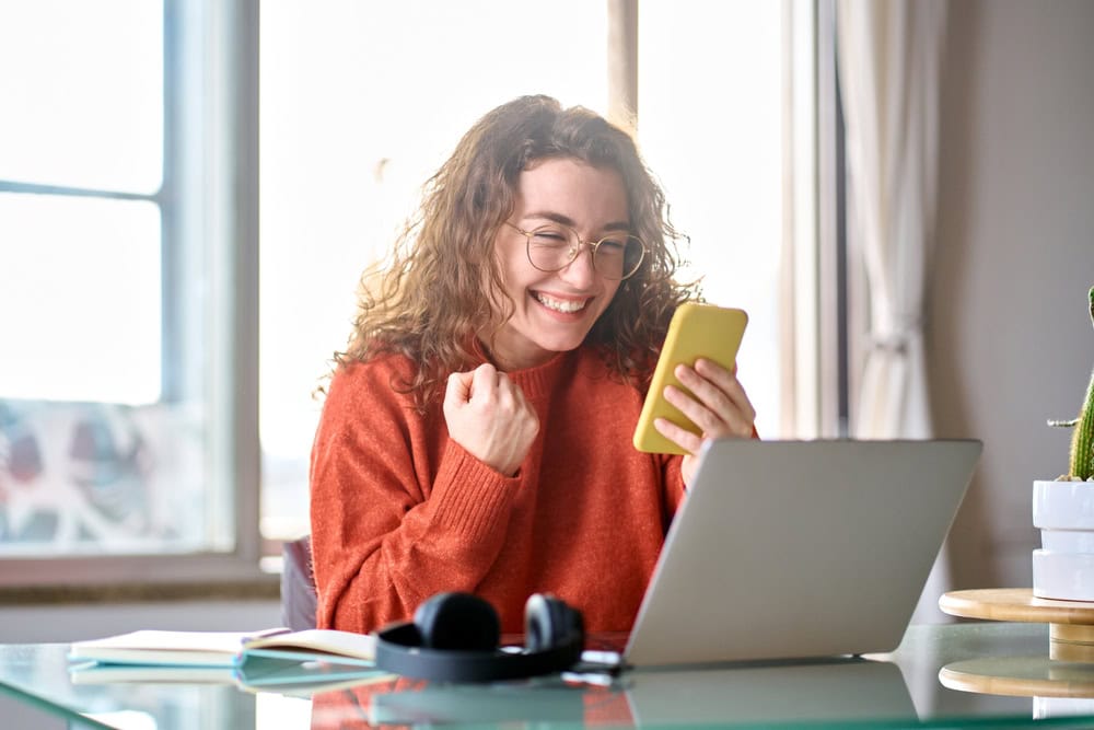 Bright woman celebrating success while working on a laptop and phone at home, coursework for becoming a licensed contractor, online learning, home office, enthusiastic about construction career.