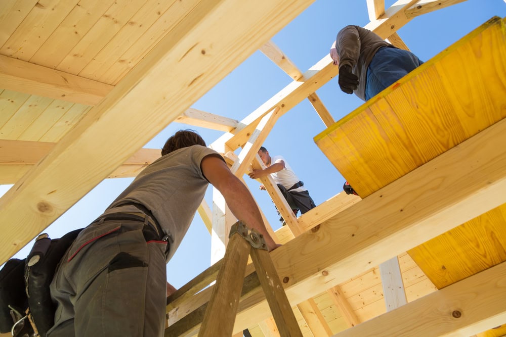 Wooden house frame construction with workers assembling the structure under clear blue sky.