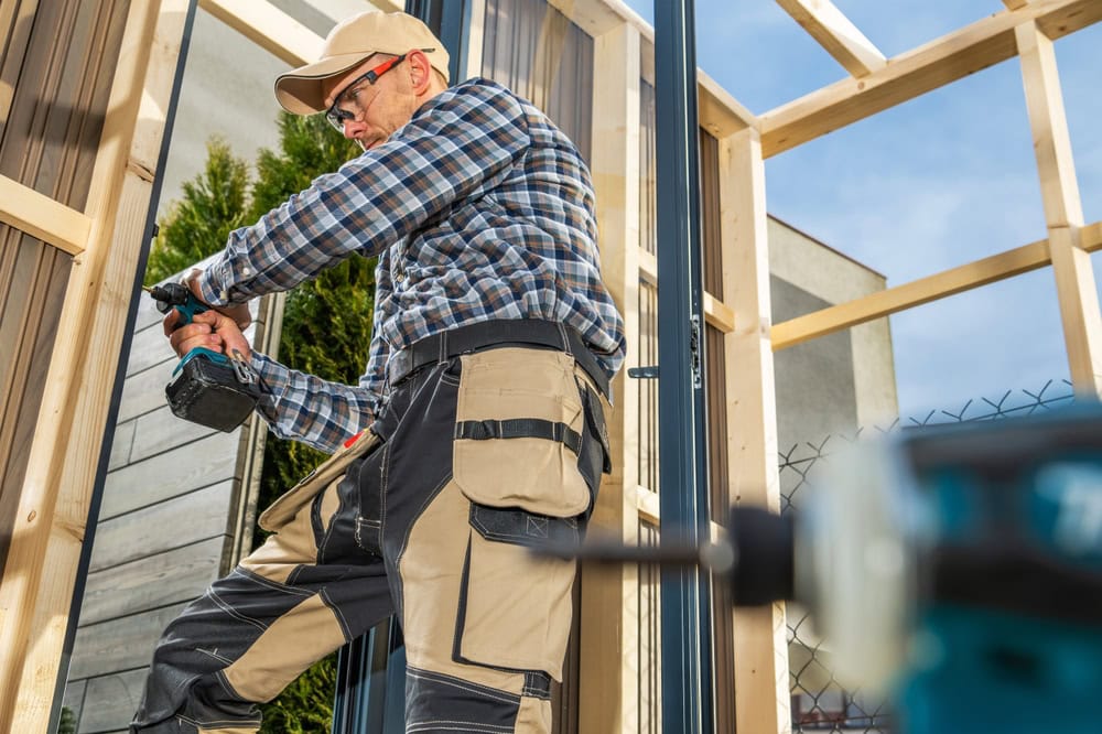 Carpenter using a cordless drill on a wooden frame at a construction site.