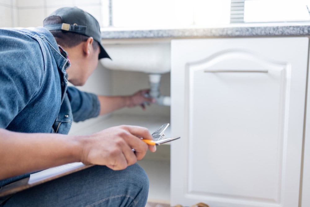 Licensed plumber inspecting under kitchen sink, checking for leaks and proper piping, professional plumbing service.