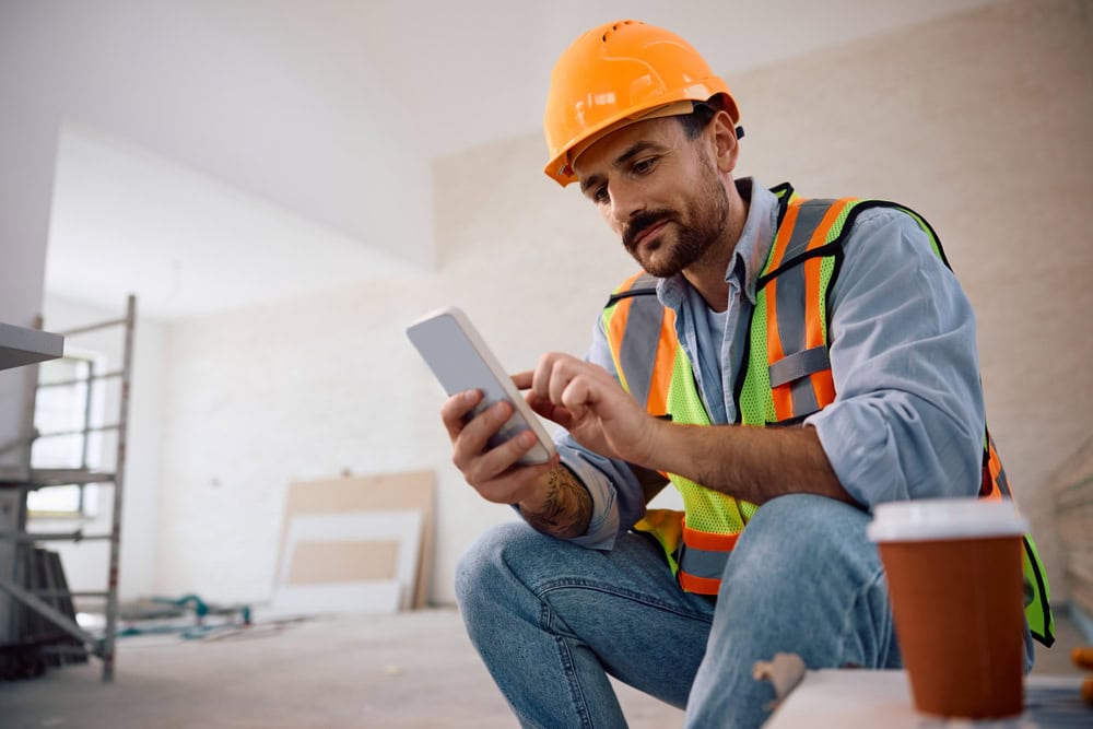 Hardhat construction worker using a tablet on an indoor job site, wearing safety vest and helmet, representing contractor training and certification.