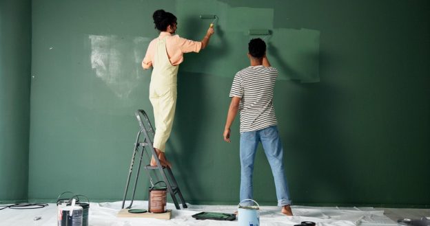 Teen girl painting wall with roller brush, helping young man in DIY home improvement project, in a room with green wall, painting supplies on floor, and ladders; interior renovation.