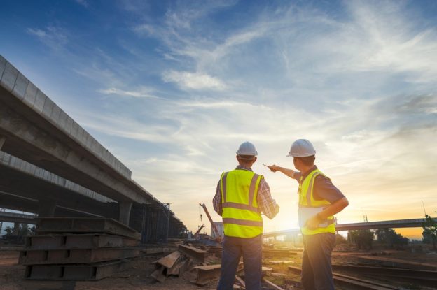 Highway construction site with two engineers in safety vests and helmets discussing at sunset, illustrating contractor training and construction project management.