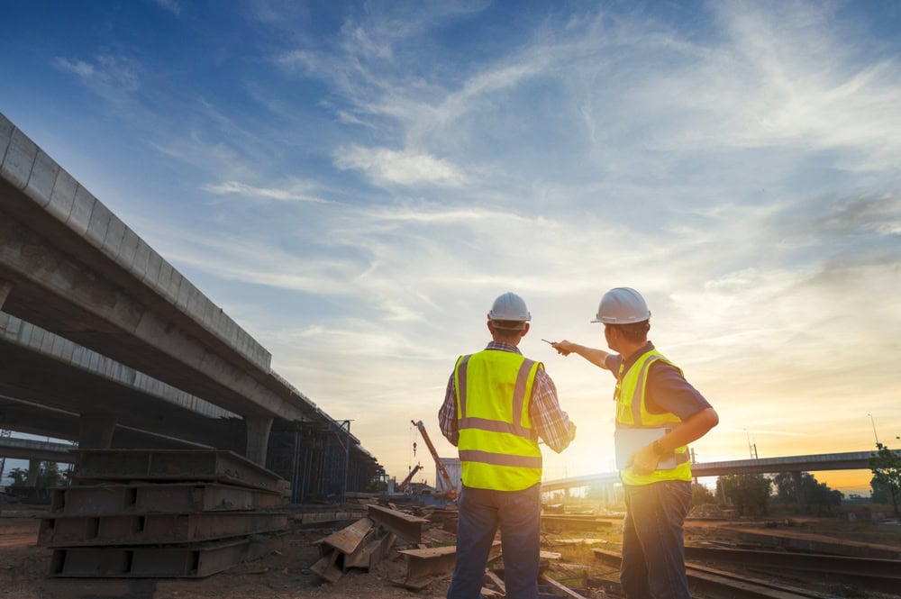 Highway construction site with two engineers in safety vests and helmets discussing at sunset, illustrating contractor training and construction project management.