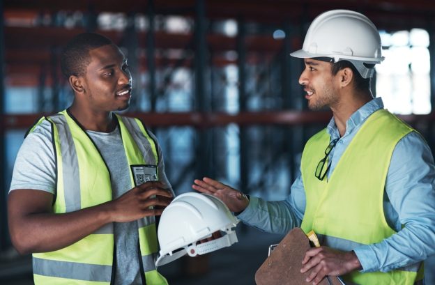 Young male construction workers discussing plans at a building site, wearing safety vests and helmets, collaborating on project details.