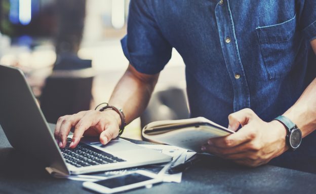 A person working on a laptop and holding a notebook, symbolizing planning and research for becoming a licensed contractor.