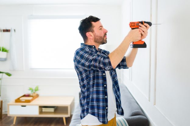 Male handyman installing a wall-mounted item with a cordless drill in a bright, modern living room.