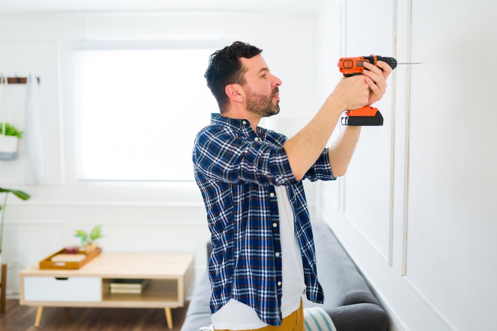 Male handyman installing a wall-mounted item with a cordless drill in a bright, modern living room.