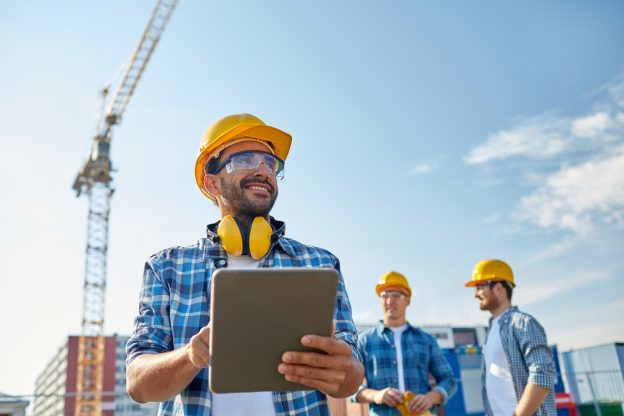 Workers wearing safety helmets and safety glasses at a construction site with cranes and buildings in the background, using a digital tablet for project management.