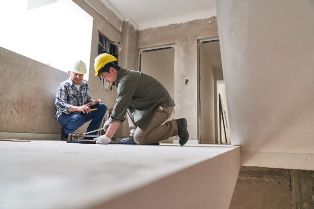 Young male construction worker measuring floor with safety helmet, working on a renovation project in an unfinished building.