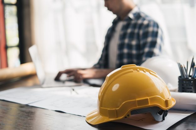Hard hat and construction plans on a desk with a contractor working on a laptop in the background.
