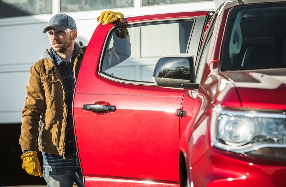 A man wearing safety gloves and glasses inspecting a red pickup truck outside a building for a contractor licensing blog.