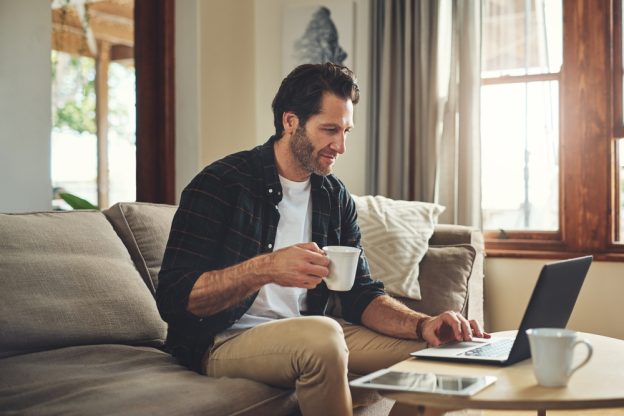 Man working on laptop in cozy home setting with coffee cup, casual smart casual attire, natural light, and warm wooden interior emphasizing professional learning at home.
