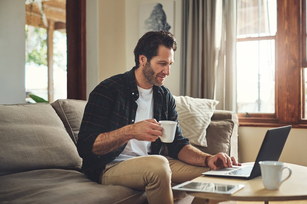 Man working on laptop in cozy home setting with coffee cup, casual smart casual attire, natural light, and warm wooden interior emphasizing professional learning at home.