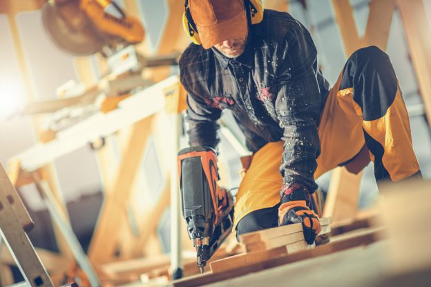 Man working with power drill in woodworking shop, wearing safety gear, engaged in carpentry task.
