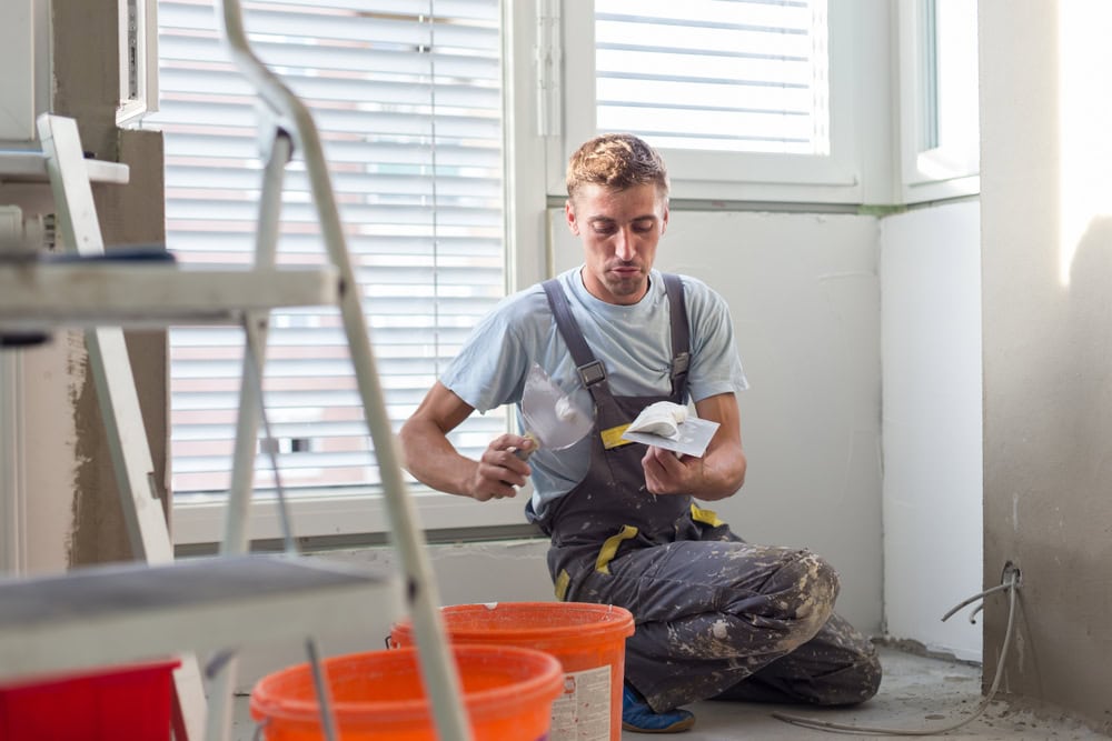 A young male construction worker reading a manual or guidebook while taking a break in a bright room with large windows, wearing work overalls and surrounded by construction tools and materials.