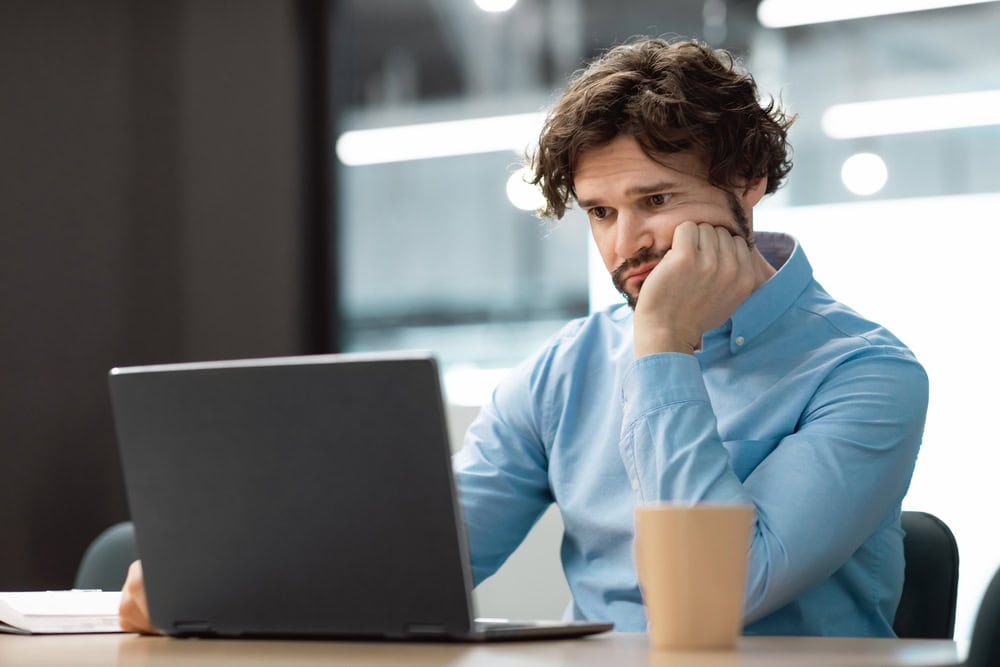 Uncomfortable man working on laptop at desk, contemplating contractor licensing or certification process at a modern office.