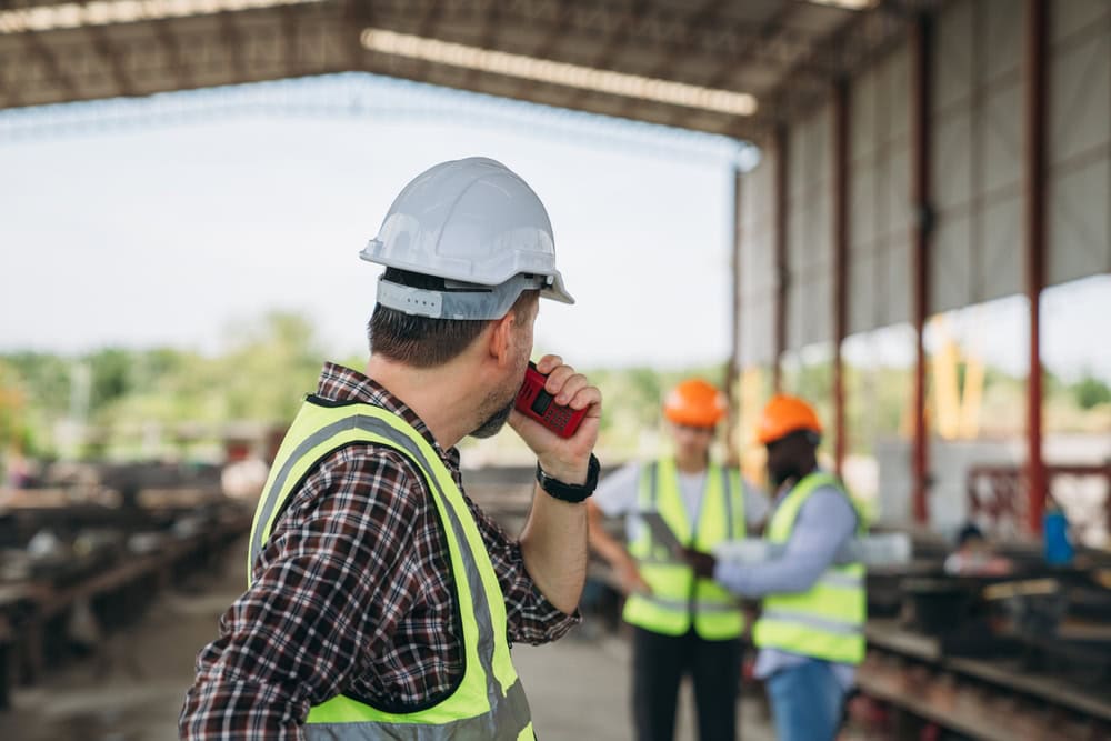 Hardhat-wearing contractor speaking on walkie-talkie at construction site with workers in safety vests in background.