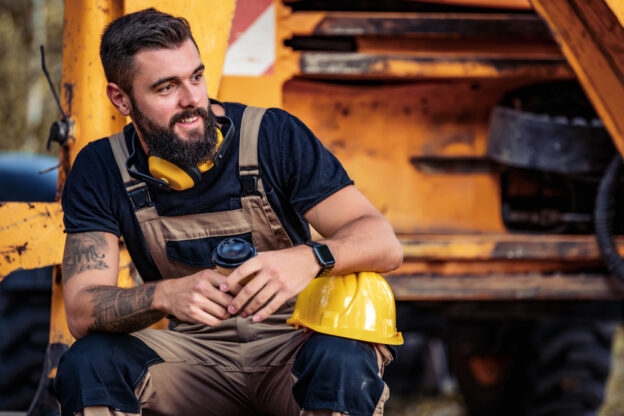 A smiling male construction worker with a beard sitting outdoors, holding a coffee cup, wearing a tool vest, with a yellow hard hat on his knee, in front of construction equipment.