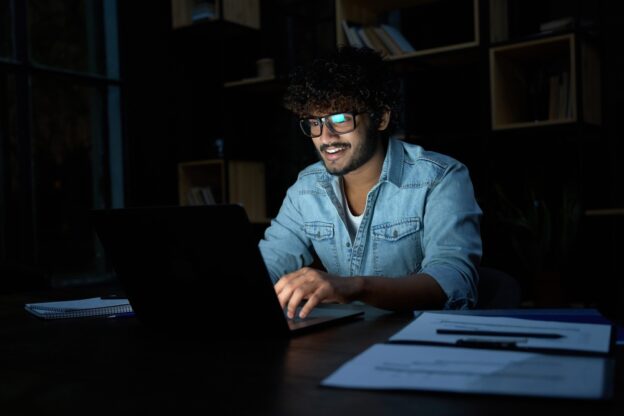 Young man working on laptop at night, studying for contractor licensing, with documents and notebooks on the desk.