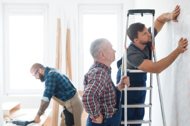 Young male contractor installing wallpaper in a bright, modern room with senior supervisor, with other team members working in background.