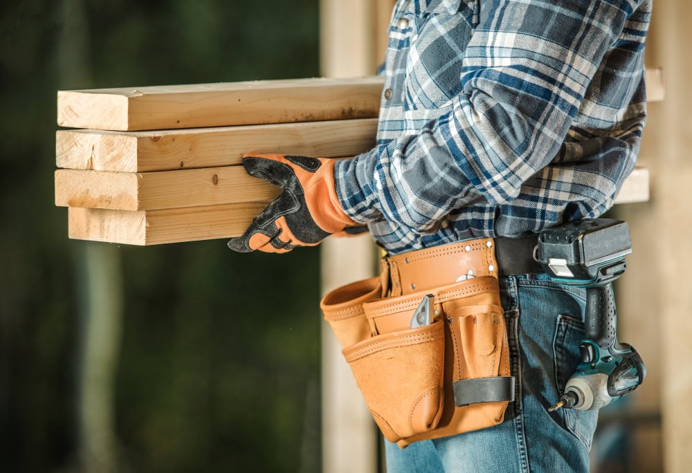 - A construction worker wearing gloves carrying wooden planks outdoors.