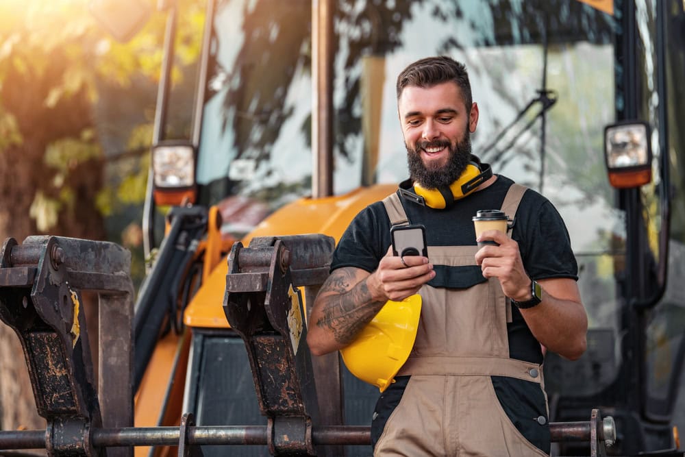Excavator operator checking phone and holding coffee at construction site with machinery in background.