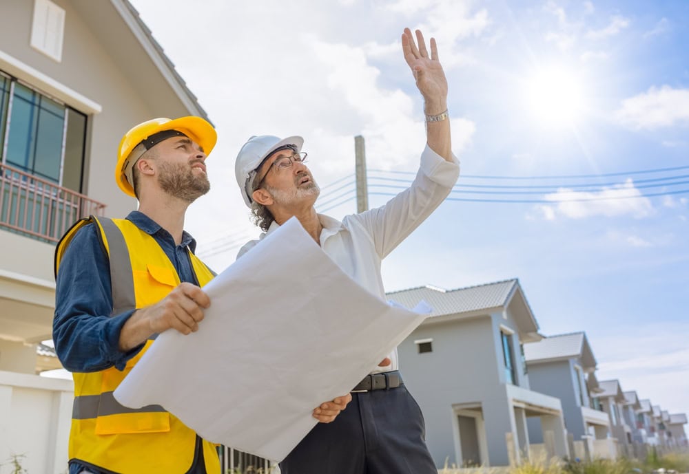 Experienced contractor guiding apprentice on construction site, reviewing blueprints under clear blue sky.