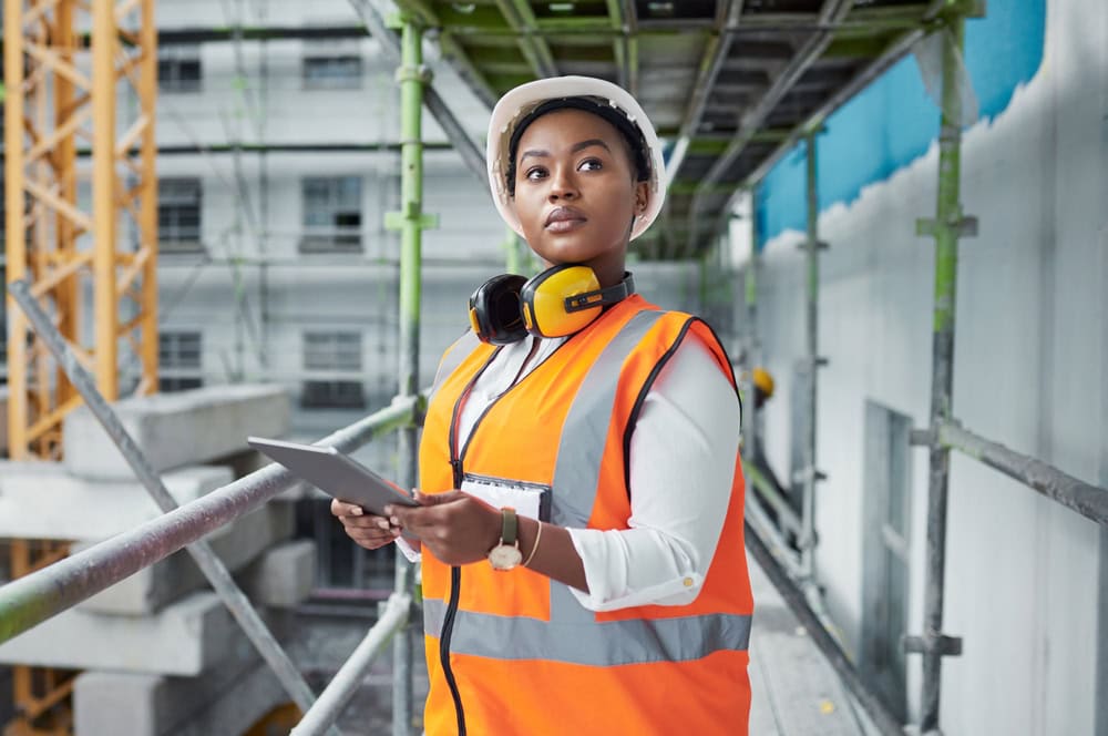Female construction supervisor inspecting site, wearing safety gear and holding digital tablet, on an active construction project.
