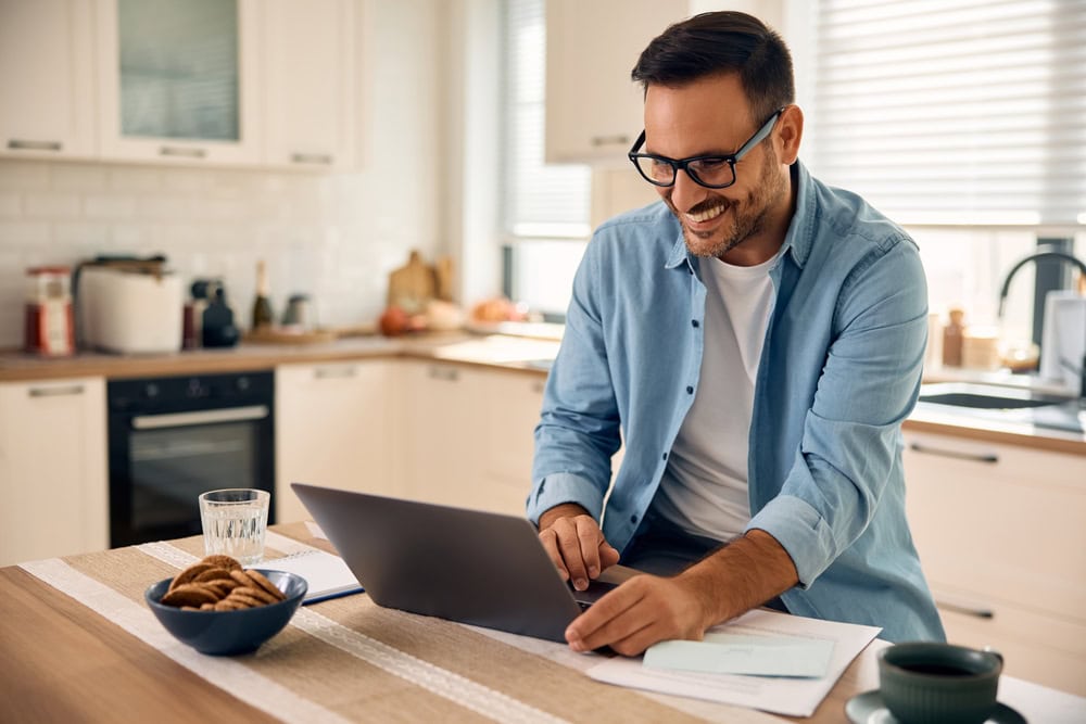 An adult man in a light blue shirt working on a laptop at a kitchen counter, smiling, with documents and cookies nearby, symbolizing contractor success and motivation.