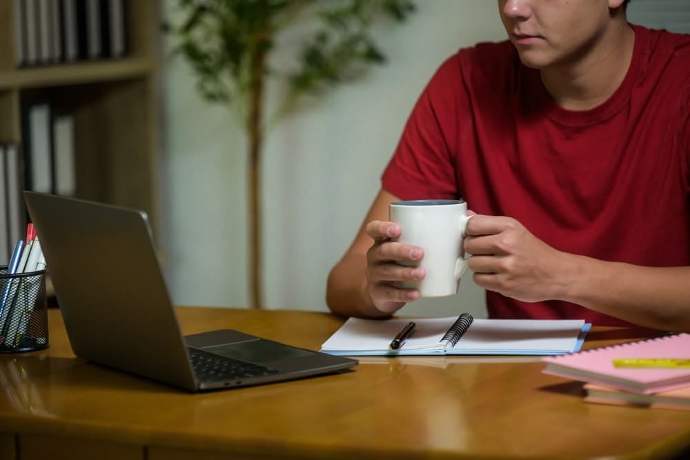 Adult man working at home on laptop, holding a coffee mug, with notebooks and pens on desk, studying or planning for contractor licensing, home office setup.