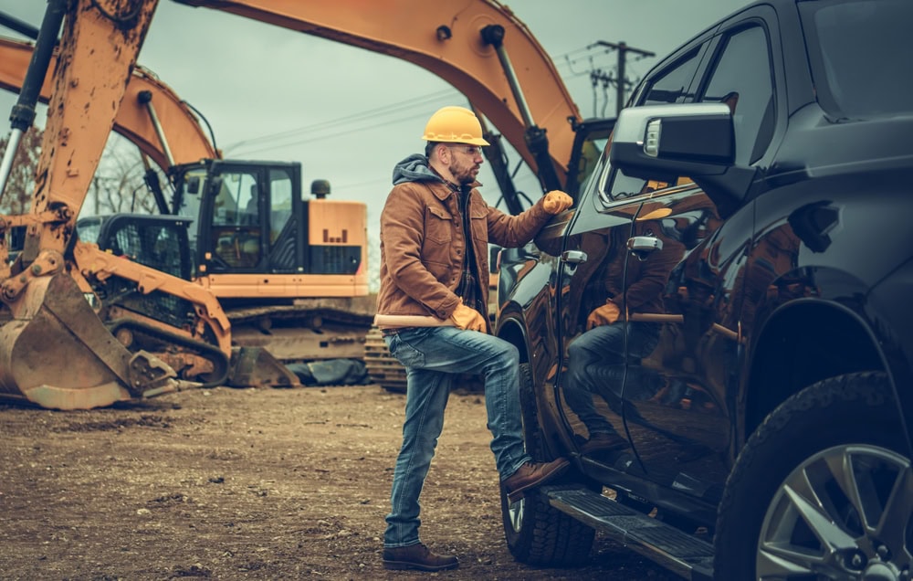 Excavator operator inspecting a black pickup truck at a construction site with heavy machinery in the background.
