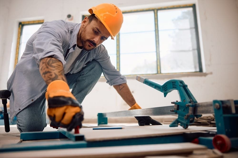 A construction worker wearing an orange safety helmet and gloves measures a wooden board with a tape measure in a well-lit indoor workspace.